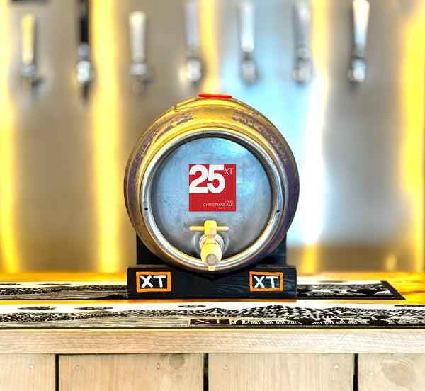 A metal 36-Pint Pin Cask of XT Brewing Co XT25 Christmas Ale on a tilt on the bar of XT Brewery's Taproom and beer pumps in the background.