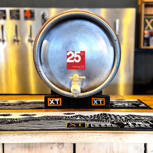 A metal Firkin 72-Pint Cask of XT Brewing Co XT25 Christmas Ale on a tilt on the bar of XT Brewery's Taproom and beer pumps in the background.